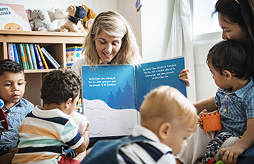 Image of two educators with a group of children sitting while reading a book