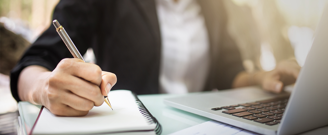 A person’s hand writing on a notebook while the other hand is using a laptop.